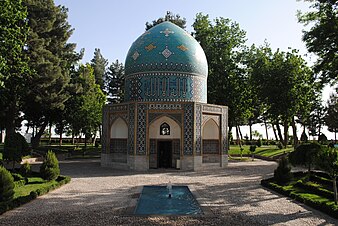 An elegant building in Islamic style, with a lower half composed of three curved-arch porches, and the top a bright blue dome, standing on a pathway in the middle of a verdant garden.