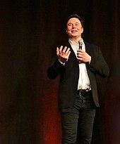 Musk speaking in front of curtains; he is holding a microphone.