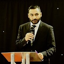Абрар Хюсейн на публично изказване в Лондон, държащ микрофон