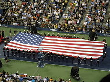 Around thiry soldiers hold a giant American flag on a blue and green hard court, surrounded by crowded bleachers