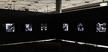 A dark space with the artist's images illuminated from within the black walls. The floor is black and the ceiling is ornate white tiles.