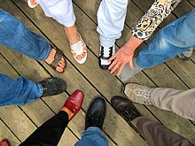 A group of people stood with one foot outstretched, forming a circle. They wear a variety of shoes and sandals.