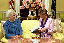 Bush and Princess Diana are seated on a couch: Diana examines an open copy of Millie's Book while Bush watches.