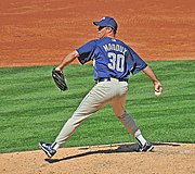 A man wearing a blue cap, blue top and light colored trousers, holding a baseball. The back of his shirt reads 