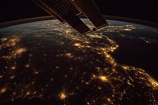 Night view of Earth from International Space Station, curve of earth intercepted by ISS solar panel in top half, city lights visiable along unknown coastline.