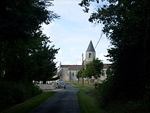 En liten trekantet vei med i bunnen en steinkirke kledd med fliser med en konisk skifertårn.  Et fartsgrenseskilt på 30 km / t til høyre.