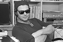 portrait noir et blanc d'un homme dans la trentaine avec les bras croisés assis au bureau devant des albums de disques vinyle et une chaîne stéréo, portant des lunettes de soleil noires, une casquette de baseball à l'envers sombre et une chemise sombre