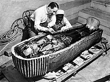 Two men examining a human-shaped coffin partly covered by black residue