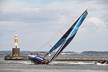 Voilier bleu gitant fortement devant une jetée au bout de laquelle se trouve un petit phare.