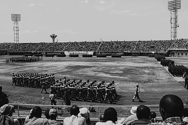 Ein unüberdachtes, auf den Rängen dicht gefülltes Stadion. Auf der Rasenfläche in der Mitte stehen Gruppen von Soldaten in rechteckigen Formationen. Weitere laufen im Gleichschritt auf der Leichtathletikbahn um diese. An den Banden des Stadions steht "Isoko Yo Kwigira", "Kwibohora 20" und "Together We Prosper".