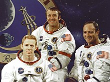 Kolorowe zdjęcie trzech członków załogi misji Apollo 14 przed logotypem i gwiaździstym tłem.