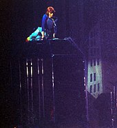 Zdjęcie mężczyzny z rudymi włosami leżącego na wybiegu w pogrążonej w ciemności scenerii, na której możemy zobaczyć sylwetkę budynku