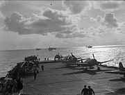 Black and white photograph of single propeller fighter aircraft on the deck of an aircraft carrier at sea. Several other ships are visible in the background.