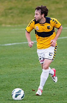 A man with dark hair in a yellow shirt with navy shorts looking up to his right with a football in front of him.