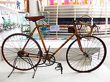 Foto av en gammel sykkel med en gul ramme plassert på et display, en hvit etikett festet til rammen.