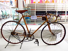 Foto van een oude fiets met een geel frame op een display, een wit label op het frame.