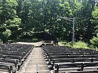 A view of the Washington Crossing Open Air Theatre in 2023. The camera is situated in the middle of the House Right seating facing the stage. The outdoor venue is showing significant signs of neglect, and part of the stage has fallen away.