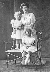 Photographie en noir et blanc d'une femme debout derrière deux petites filles blondes.