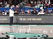 Sandy Brondello coaching for the NY Liberty in August, 2024. She stands along the courtside, where the advertising behind her reads "The Ball is in Your Court." Liberty Mascot Ellie the Elephant is draped over the stands in the crod behind her