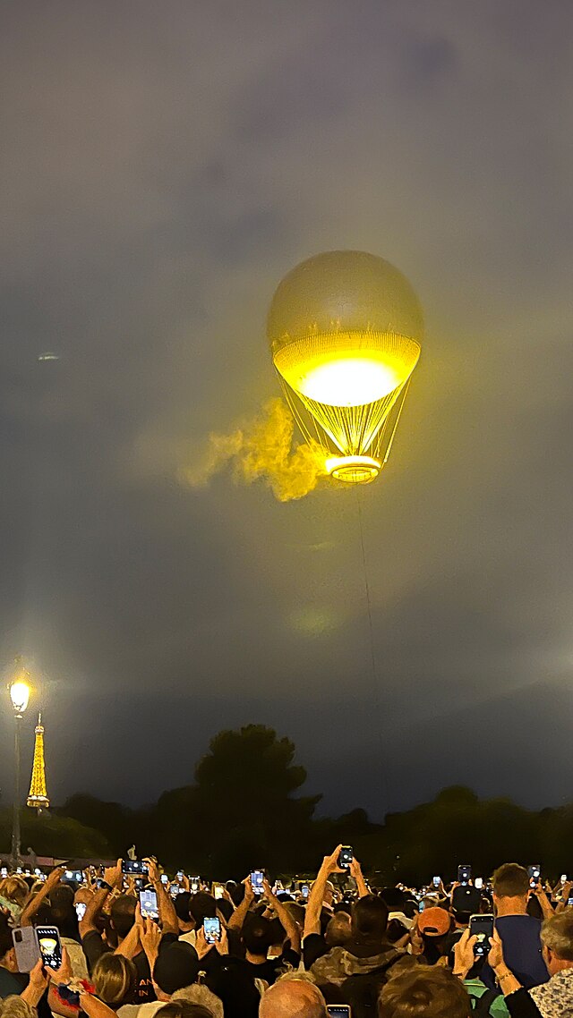 Ein Nachtbild mit aufgestiegenem gefesselten Ballon, der eine Feuerschale trägt. Am Horizont links unten der beleuchtete Eiffelturm.