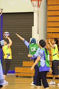 Erwachsene Frauen aus Brunei spielen Netball.Ein Team trägt grün und das andere Team trägt gelb.Das Spiel wird drinnen auf Holzböden gespielt.Die Spieler tragen alle Hosen.Einige haben Bedeckungen über ihren Köpfen.Das gelbe Team schießt gerade und das grüne Team versucht, den Schuss zu blockieren.