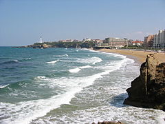 Vue d'une plage sur laquelle les vagues se brisent ; phare et bâtiments en arrière-plan.