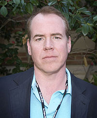 Bret Easton Ellis is pictured standing on front of a stone wall and greenery. He is wearing a blazer over a blue collared shirt and has a lanyard on his neck.