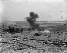 A black-and-white photograph of a rocky landscape with a puff of smoke from a dynamite explosion