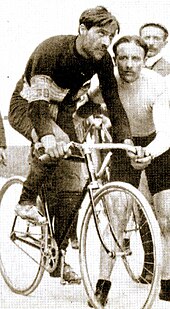 Sort og hvidt fotografi af en mustached mand, klædt i sort, sidder på sin cykel og omgivet af to andre mænd.
