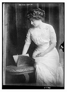 A young white woman, seated, wearing a white gown with short sleeves; one hand in her lap; she is gazing down at an open soft-cover book on the stool next to her