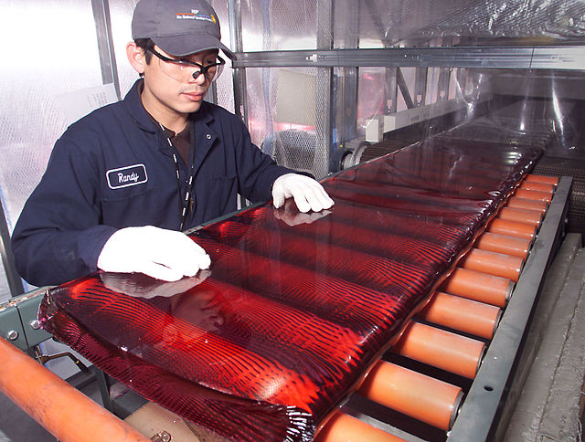 Un ouvrier portant des gants et une casquette penché sur un long bloc rouge translucide roulant sur une chaîne de fabrication.