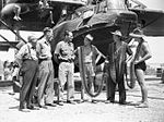 Members of 20 Squadron with lines of machine gun belts wrapped around them speak with members of the parliamentary war expenditure committee.