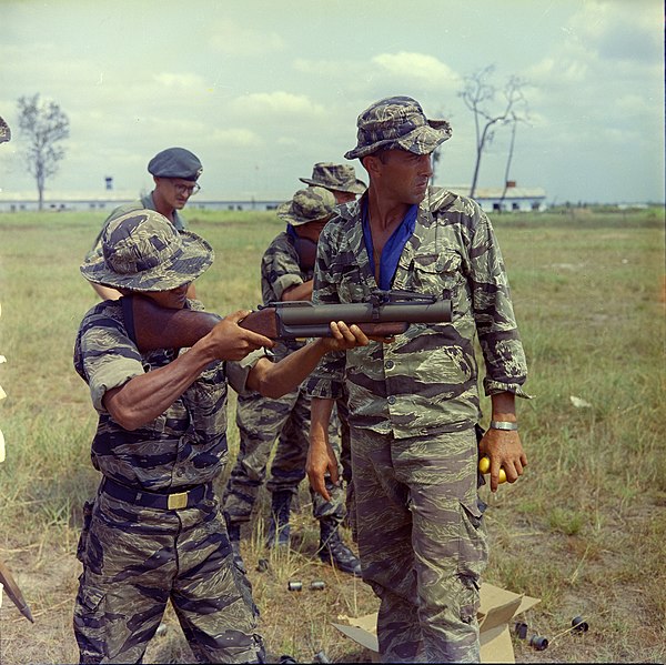 A United States Army Special Forces advisor instructing a Vietnamese Civilian Irregular Defense Group trainee on how to use an M79 grenade launcher