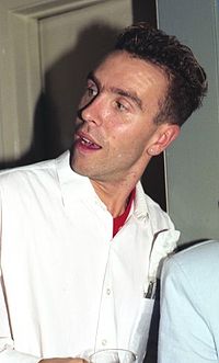 Upper body shot of a 28-year-old man, his head is twisted to face his right. His mouth is open. He has dark, short, curly hair and wears a white shirt. Behind him is the room's closed door with the shadow of his head cast onto it.