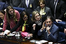 Da esquerda para a direita, encontra-se Vanessa Grazziotin, apoiada sob uma Mesa do Senado. Do lado dela, Fátima Bezerra, Gleisi Hoffmann e Eunício Oliveira, este último falando ao microfone. Estão sentados em frente a uma mesa. Atrás deles, há um aglomerado de pessoas