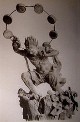 Three-quarter view of a statue. His left leg is bend as if climbing stairs and he is leaning forward. His hair is standing up and his right hand is raised above his head. A thin halo around his head is decorated with thick circular disks. Black and white photograph.