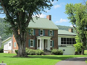 An image of two-story brick house with a green roof, a large front porch, and a tree in the foreground.