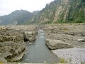 Bir derenin (ortada) kesiştiği bir kanyonun katmanlı bej kayalıklarının renkli fotoğrafı.  Sütlü bir gökyüzü altında arka planda dağlar.