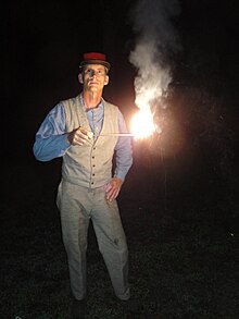 A man is standing in the dark. He is holding out a short stick at mid-chest level. The end of the stick is alight, burning very brightly, and emitting smoke.