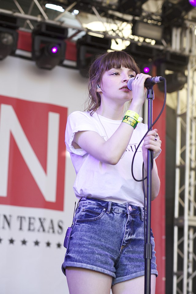 A young woman with brown hair, wearing a white T-shirt and denim shorts, singing into a microphone