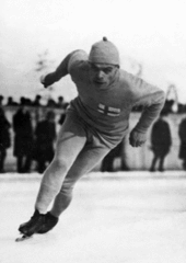 Czarno-białe zdjęcie mężczyzny jeżdżącego na łyżwach szybkich, pochylonego do przodu, lekko ugiętych nóg, cofniętych ramion, noszącego fińską flagę na piersi.