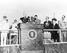 A black and white photograph of President Harry Truman standing at a podium bearing the presidential seal on a stage with people behind him applauding