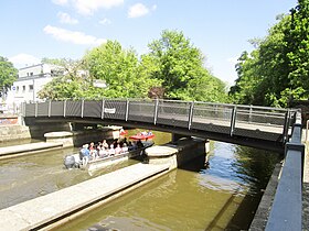 Steel foot bridge over water against a blue sky with green trees