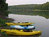 One blue and two yellow kayaks on the shore of a lake, which reflects surrounding pine trees