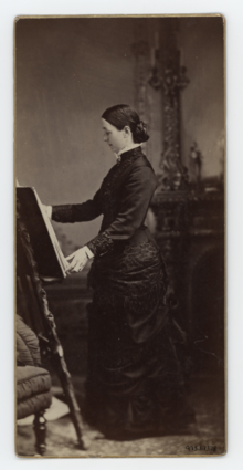 A black and white photograph of a woman in a dark dress standing in profile at an easel.