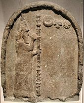 Stenstele med en udskæring, der skildrer en mand med et skæg, bærer en høj stav og iført en kappe og keglehue, der gestikulerer til tre symboler, der repræsenterer månen, solen og Venus.