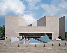 A large grey building rises above a stone plaza. Short square towers appear on either side of the building, and an array of irregular glass pyramids are in the middle of the plaza.