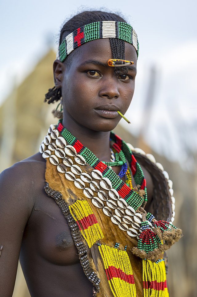 אישה משבט האמר; היא עומדת חצי ערומה, סביב צוואה תכשיטים רבים עשויים חרוזים וצדפות. שדה השמאלי נראה לצד השרשראות. על מצחה בנדנה עשויה חרוזים.