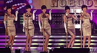 Five women with beehive hairstyles wearing matching golden sheath dresses and elbow-length gloves