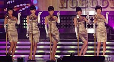 Five women with beehive hairstyles wearing matching golden sheath dresses and elbow-length gloves